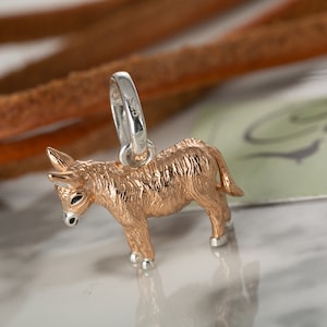 Op de afbeelding: Een kleine, gouden ezelbedel met een zilveren lus om aan een armband of ketting te bevestigen. De ezel staat op alle vier poten en heeft een gedetailleerd, getextureerd oppervlak.