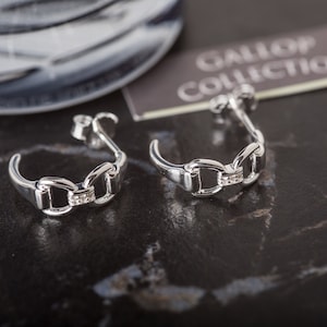 Peut inclure: Une paire de boucles d'oreilles créoles en argent avec un design de chaîne et de petites pierres claires. Les boucles d'oreilles sont sur une surface gris foncé avec une étiquette qui dit "GALLOP COLLECTION".