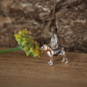 May include: A small, three-dimensional dog charm crafted from polished silver with rose gold accents on the head and ears. The charm features a circular jump ring at the top for attachment, depicting a standing dog with a smooth, metallic finish.