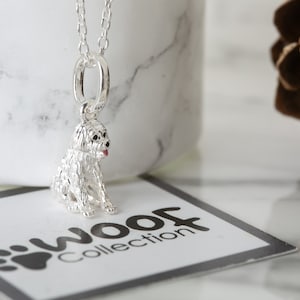 May include: Silver charm necklace featuring a detailed, white, fluffy dog charm with a pink tongue. The charm is hanging from a silver chain and is sitting on a white surface with a black and white card that says "WOOF Collection".