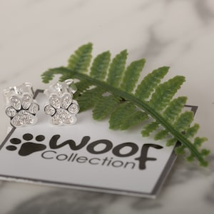 May include: A pair of silver paw print earrings with clear crystals. The earrings are on a white background with a small card that says "woof collection".