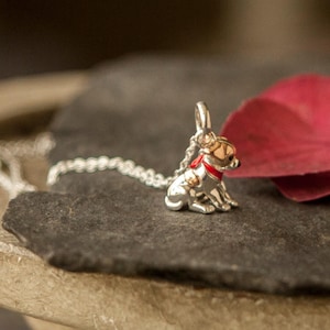 Op de afbeelding: Een zilverkleurige metalen bedel in de vorm van een kleine zittende hond, voorzien van een goudkleurige kop, een felrode halsband en een dunne zilveren ketting bevestigd aan een ringetje aan de bovenkant, bedoeld als kettinghanger.