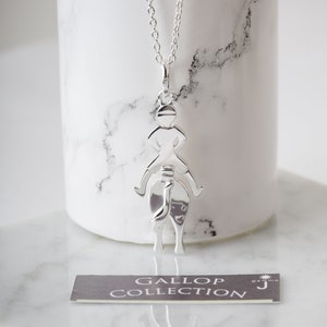 May include: A silver necklace with a pendant of a jockey riding a horse. The pendant is a silhouette of a jockey and horse. The necklace is on a white marble surface. The text "GALLOP COLLECTION" is visible on a small card in the image.