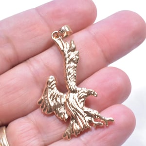May include: A gold-colored pendant in the shape of a bird, possibly an eagle or hawk, with outstretched wings. The pendant has intricate details and is held in a hand against a white background. The bird is facing to the right.