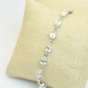 May include: A silver bracelet with clear crystal beads. The bracelet is on a beige fabric surface.
