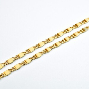 K&ouml;nnte beinhalten: Nahaufnahme von zwei goldfarbenen Halsketten. Die Ketten haben ein zartes, flaches, oval geformtes Kettenglied-Design. Der Schmuck ist vor einem schlichten wei&szlig;en Hintergrund platziert, der die helle, reflektierende Oberfl&auml;che des Metalls hervorhebt.