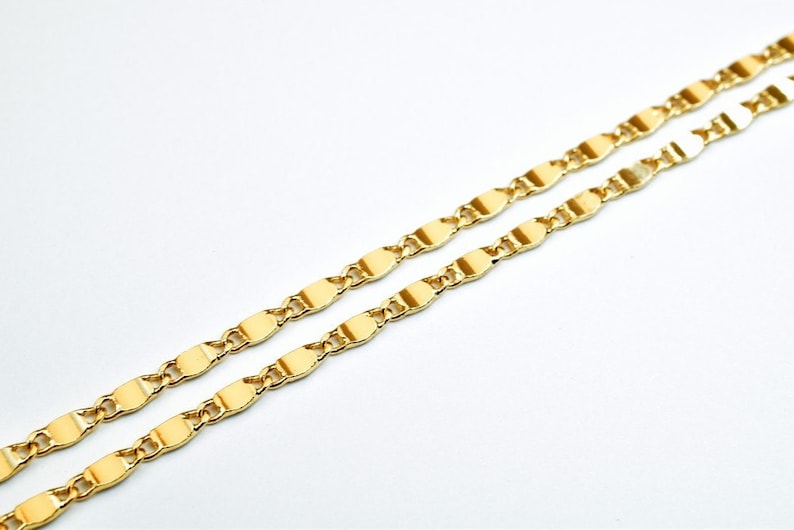 K&ouml;nnte beinhalten: Nahaufnahme von zwei goldfarbenen Halsketten. Die Ketten haben ein zartes, flaches, oval geformtes Kettenglied-Design. Der Schmuck ist vor einem schlichten wei&szlig;en Hintergrund platziert, der die helle, reflektierende Oberfl&auml;che des Metalls hervorhebt.