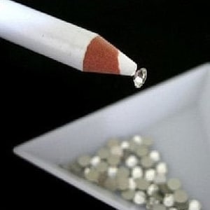 May include: A white pencil with an orange tip is holding a small, clear rhinestone above a white, square tray. The tray contains a pile of small, silver rhinestones. The background is black.
