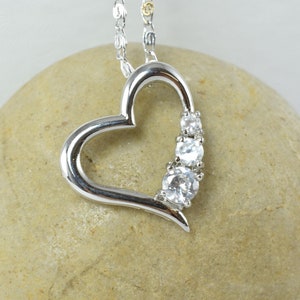May include: A silver heart-shaped pendant necklace with a delicate chain. The pendant features a row of three clear, round gemstones. The necklace is displayed on a light brown stone against a white background.