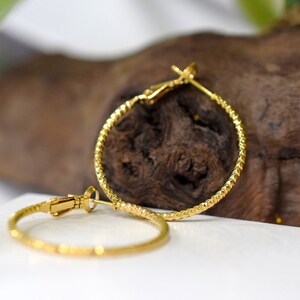 May include: A pair of gold hoop earrings with a textured design. The earrings are on a wooden surface.