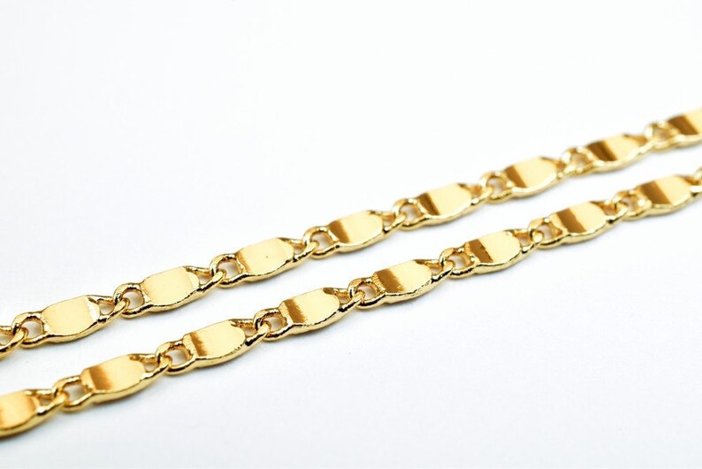 K&ouml;nnte beinhalten: Nahaufnahme einer goldfarbenen Halskette. Die Kette besteht aus flachen, oval geformten Gliedern, die durch kleine, runde Glieder verbunden sind. Die Halskette wird vor einem wei&szlig;en Hintergrund pr&auml;sentiert, der ihre gl&auml;nzende, reflektierende Oberfl&auml;che hervorhebt.