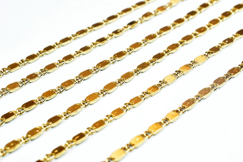 May include: Close-up of multiple gold-colored chain necklaces. Each chain features a series of elongated, oval-shaped links connected by smaller, round links. The chains are arranged diagonally against a white background.