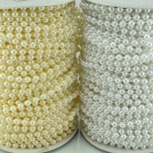 May include: Two spools of beaded trim. One spool is a creamy ivory color, and the other is white. The beads are small and round, strung together to create a continuous strand. The spools sit on a white base.