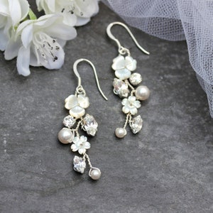 May include: A pair of silver dangle earrings with white pearl and crystal accents. The earrings feature a cluster of small white flowers and a larger white flower with a pearl centre.