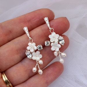 May include: Elegant silver earrings with white flower details, sparkling crystals, and pearl drops. These dangle earrings are ideal for weddings or special events. The earrings are displayed against a soft white backdrop.