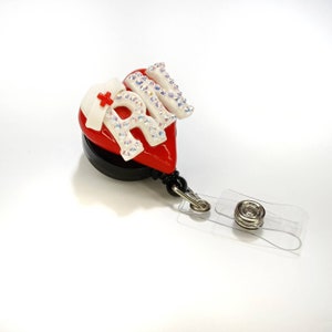 Pode incluir: Porta-crachá retrátil em forma de coração vermelho com a palavra "RN" em letras brancas com strass. Um gorro de enfermeira branco com uma cruz vermelha está no coração.