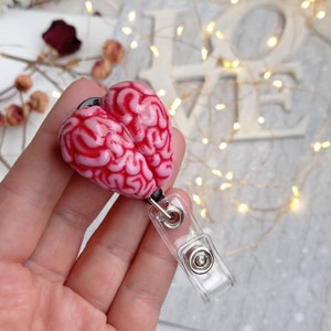 Pode incluir: Um chaveiro em forma de cérebro rosa e vermelho com um porta-crachá retrátil de plástico transparente.