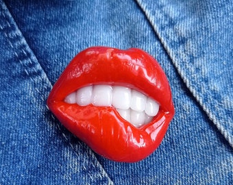 Puede incluir: Una escultura de labios de resina roja y brillante con dientes blancos mordiendo el labio inferior. La escultura está sobre un fondo de mezclilla azul.