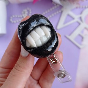 Pu&ograve; includere: Un portabadge a forma di bocca di vampiro in bianco e nero. La bocca &egrave; realizzata in pasta polimerica e ha denti bianchi affilati. Il portabadge &egrave; attaccato a un cinturino in plastica trasparente.