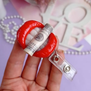 May include: Red lips with white teeth holding a rolled up dollar bill with the text "1599" visible. The lips are attached to a clear plastic badge holder.