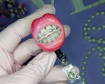 Puede incluir: Labios de plástico rosa con brackets plateados y un portagafete retráctil de plástico transparente.