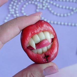 May include: A red, open mouth with sharp, white fangs and a pink tongue. The mouth has a black cross drawn on the bottom lip.