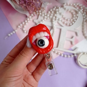 Pode incluir: Um porta-crach&aacute; de pl&aacute;stico vermelho e branco com uma boca e um olho de desenho animado. A boca est&aacute; aberta e tem dentes brancos. O olho &eacute; branco com uma pupila preta.