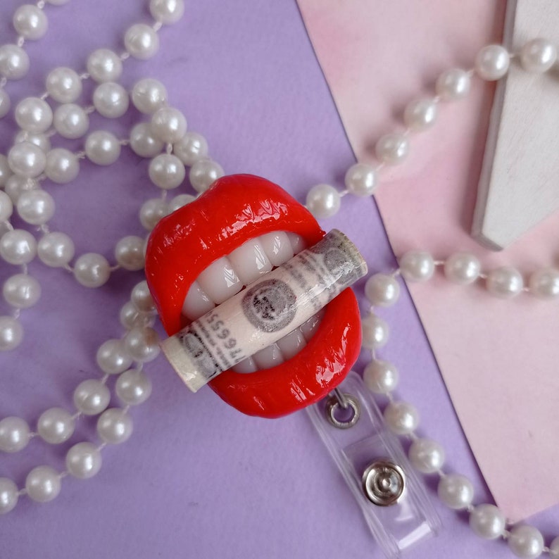 May include: Red lips with white teeth holding a rolled up dollar bill, attached to a white pearl necklace and a clear plastic badge holder.