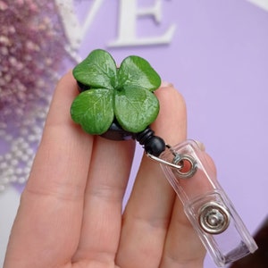 Puede incluir: Un portagafete retr&aacute;ctil negro con un amuleto de tr&eacute;bol de cuatro hojas verde y un clip de pl&aacute;stico transparente.