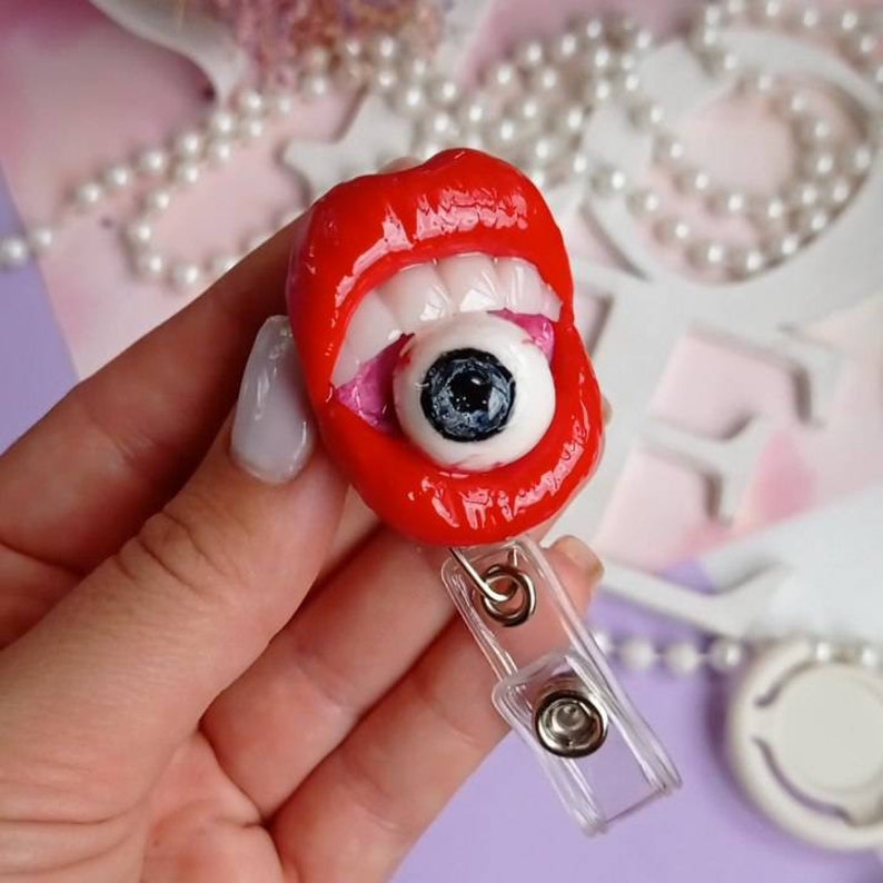 Pode incluir: Uma boca vermelha aberta com dentes brancos e um globo ocular grande, preto e branco dentro. A boca est&aacute; presa a um porta-crach&aacute; de pl&aacute;stico transparente.