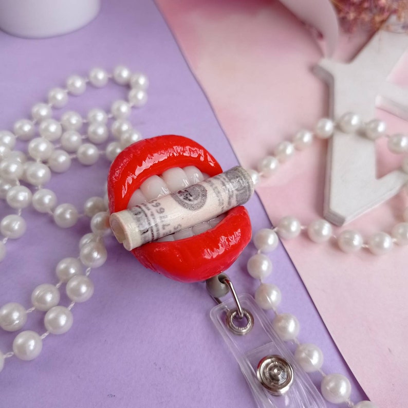 May include: A red, sculpted mouth with white teeth holding a rolled-up dollar bill. The mouth is attached to a white pearl necklace with a clear plastic badge holder.