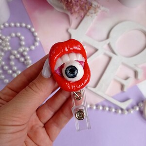 Pode incluir: L&aacute;bios vermelhos com dentes brancos segurando um globo ocular grande com uma pupila preta. Os l&aacute;bios est&atilde;o presos a um porta-crach&aacute; retr&aacute;til de pl&aacute;stico transparente.