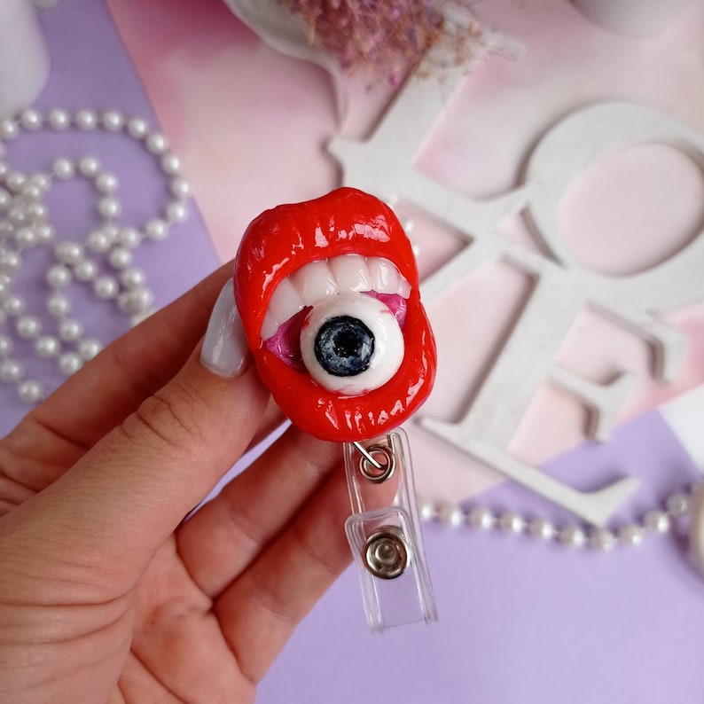 Pode incluir: L&aacute;bios vermelhos com dentes brancos segurando um globo ocular grande com uma pupila preta. Os l&aacute;bios est&atilde;o presos a um porta-crach&aacute; retr&aacute;til de pl&aacute;stico transparente.