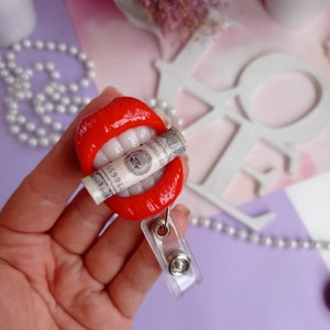 May include: Red lips with white teeth holding a rolled up dollar bill. The lips are attached to a clear plastic retractable badge holder.