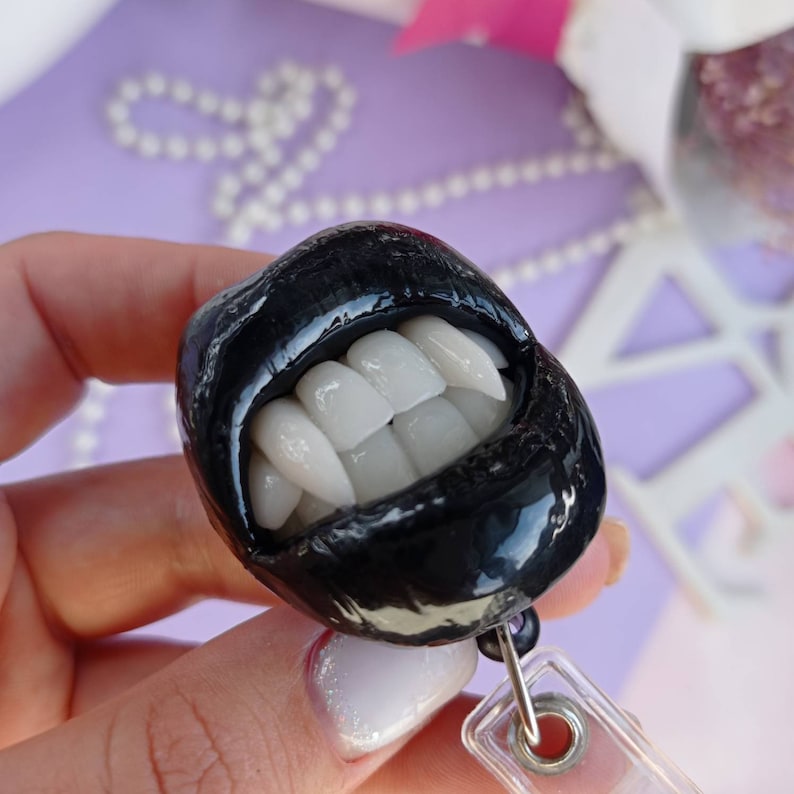 Pu&ograve; includere: Ciondolo in resina nero e bianco di una bocca con zanne, attaccato a un porta badge in plastica trasparente con una clip in metallo argentato.