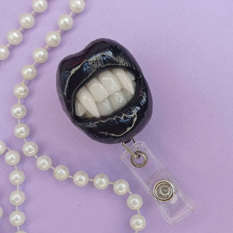 Pu&ograve; includere: Ciondolo a forma di bocca con denti di vampiro in bianco e nero su una catena di perle bianche con un porta badge retrattile in plastica trasparente.
