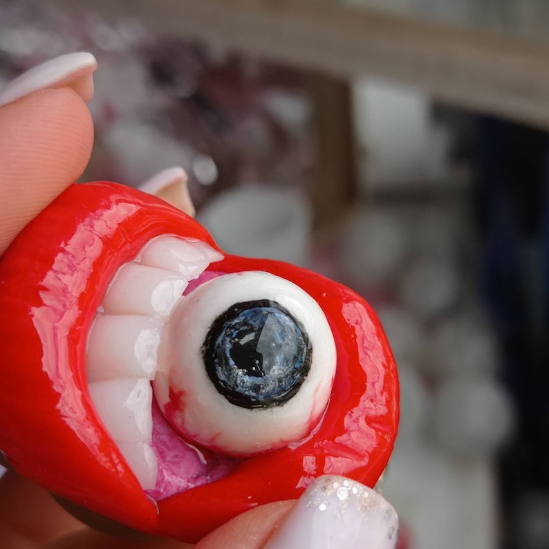 Pode incluir: Uma boca vermelha aberta com dentes brancos e um globo ocular branco grande com uma pupila preta e uma &iacute;ris azul. O globo ocular est&aacute; parcialmente dentro da boca.