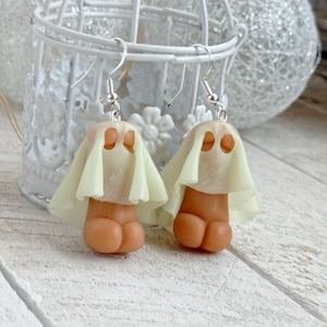 May include: A pair of dangle earrings featuring a ghost-like figure with a white veil and a brown body. The earrings are made of polymer clay and have silver hooks.