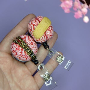 May include: Two pink and white brain-shaped keychains with a zipper and a bandage on them. The keychains are attached to clear plastic retractable badge holders.