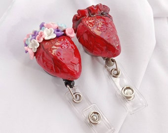 Puede incluir: Dos encantos de resina en forma de corazón anatómico rojo con acentos florales. Un corazón tiene una corona floral de flores rosas, blancas y moradas. Ambos corazones están unidos a carretes de insignia retráctiles.