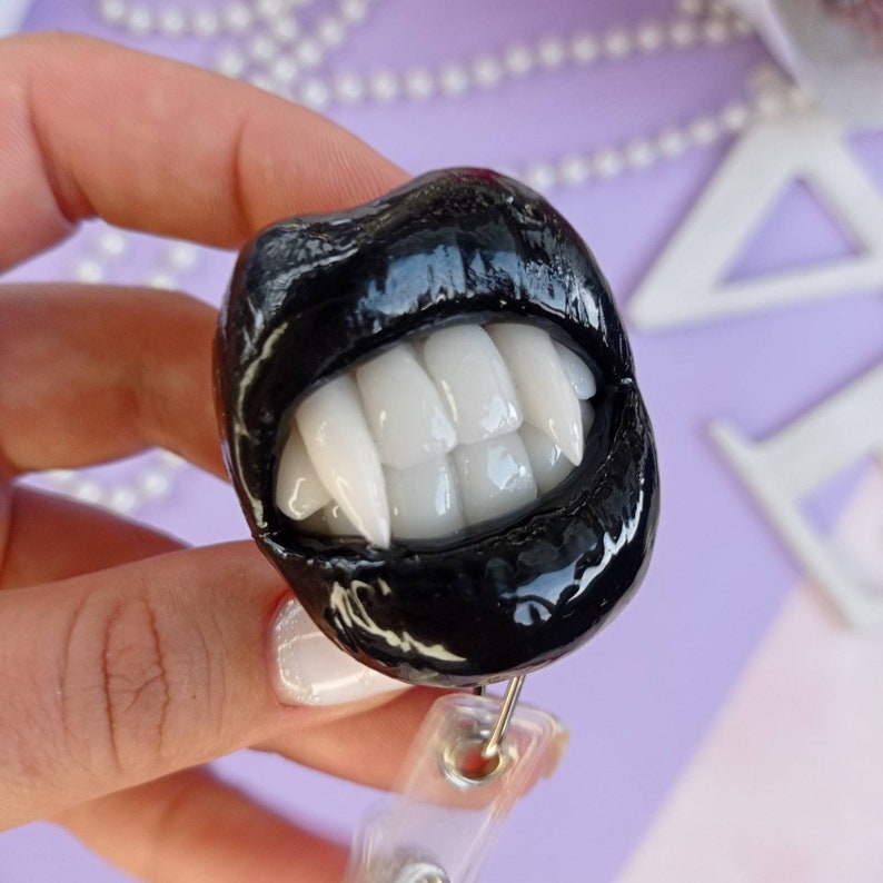 Pu&ograve; includere: Un ciondolo a forma di denti di vampiro bianco e nero con una finitura nera lucida. Il ciondolo &egrave; attaccato a un porta badge retrattile in plastica trasparente.