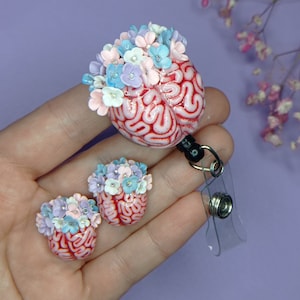 Pode incluir: Um cérebro rosa e vermelho com uma coroa de flores rosa pastel, azul e branco. O cérebro está preso a um porta-crachá retrátil com uma alça de plástico transparente.