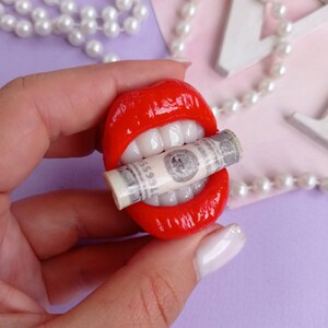 May include: Red lips with white teeth holding a rolled up dollar bill. The lips are made of polymer clay.