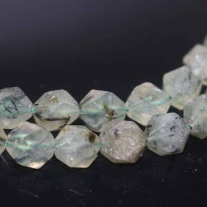 May include: A strand of faceted, translucent green and black gemstone beads. The beads are irregularly shaped and have a rough, natural texture.
