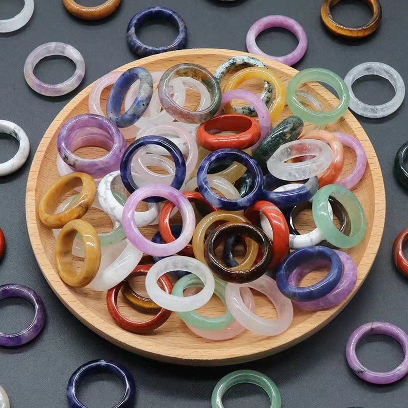 May include: A collection of colorful gemstone rings in various shapes and sizes. The rings are made of natural stones and are arranged in a wooden bowl.