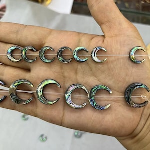 Pode incluir: Um fio de contas de meia lua de madrepérola de abalone iridescente. As contas são enfiadas em um cordão elástico transparente e estão prontas para serem usadas na fabricação de joias.