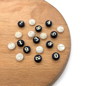 May include: A collection of black and white round beads with gold numbers 0 through 9. The beads are arranged on a wooden surface.