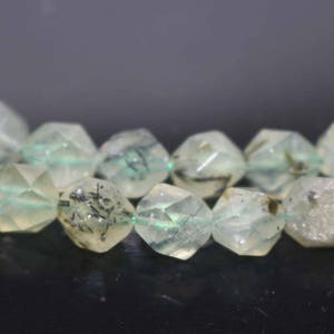 May include: A strand of faceted green and white gemstone beads with black inclusions. The beads are strung on a clear thread.