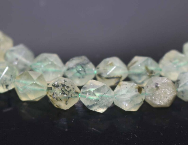 May include: A strand of faceted green and white gemstone beads with black inclusions. The beads are strung on a clear thread.
