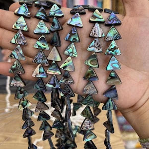 Puede incluir: Una hebra de cuentas triangulares de concha de abulón iridiscente. Las cuentas son una mezcla de colores verde, azul y marrón. Las cuentas están enhebradas en un hilo negro.